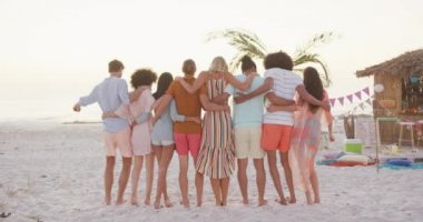 Günbatımında tropikal bir plajda eğlenen, kucaklaşan, dans eden ve ağır çekimde el kaldıran çok ırklı arkadaş grubunun arka görüntüsü.