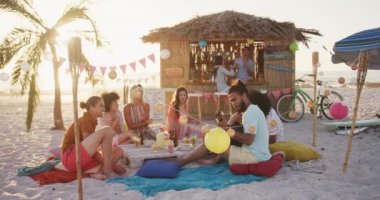 Gün batımında tropikal bir plajda oturup ağır çekimde gülerek eğlenen melez ırk arkadaşlarının ön görüntüsü.