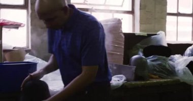 Bir şapka fabrikasındaki atölyede çalışan orta yaşlı melez bir adamın arka planda buhar ve ekipmanla şapka şekillendirip esnetirken yan görüntüsü.