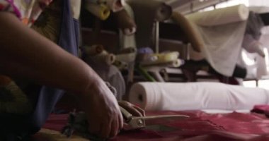 Ön, görüş, melez, ırk, kadın, giysi, önlük, pürüzsüz yan görünüm. Önlük giymiş, şapka fabrikasının atölyesinde bir parça malzeme kesen, arka planda malzeme ve ekipman bulunan, yavaş çekte, melez bir kadının görüntüsü.