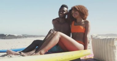 Afrika kökenli Amerikalı bir çiftin, tropik bir sahilde güneşin tadını çıkarırken, battaniyede oturup yavaş çekimde bir şeyi kucaklarken ve işaret ederken görüntüsü.