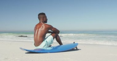 Afrika kökenli Amerikalı bir adamın tropikal bir plajda güneşin tadını çıkarırken mavi sörf tahtasının yanında yavaşça otururken görüntüsü.