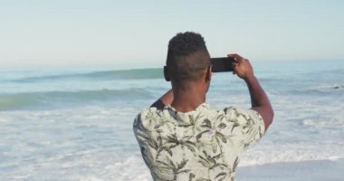 Afrika kökenli Amerikalı bir adamın tropik bir plajda güneşin tadını çıkarırken yavaş çekimde denizin panoramik bir fotoğrafını çekerken görüntüsü.