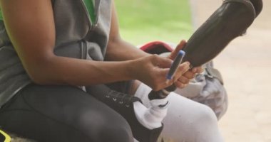 Engelli bir melez adamın yan görüntüsü. Parkta çalışıyor, duvarda oturuyor, protez bacağını tamir ediyor, kulaklıkla, ağır çekimde müzik dinliyor.