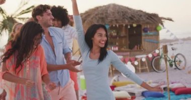 Gün batımında tropik bir plajda eğlenen, gülümseyen, dans eden ve ağır çekimde el kaldıran çok ırklı arkadaş grubunun ön görüntüsü.