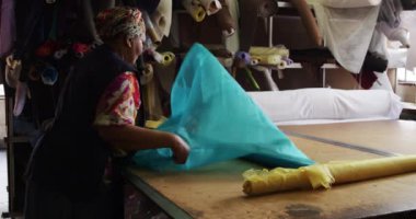 Bir şapka fabrikasının atölyesinde önlük giymiş, üzerinde malzeme ve ekipman bulunan bir kumaşı katlayan ve ölçen melez bir kadının yan görüntüsü.