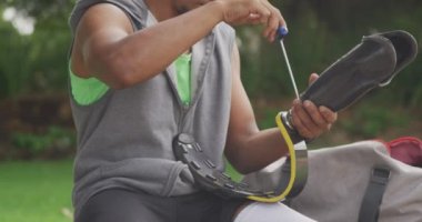 Engelli bir melez adamın yan görüntüsü. Parkta çalışıyor, duvarda oturuyor, protez bacağını tamir ediyor, kulaklıkla, ağır çekimde müzik dinliyor.