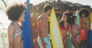 Çok ırklı bir arkadaş grubunun, tropikal bir plajda güneşlenip sörf tahtalarını tutarak yavaş çekimde gülümsemesinin ön görüntüsü.