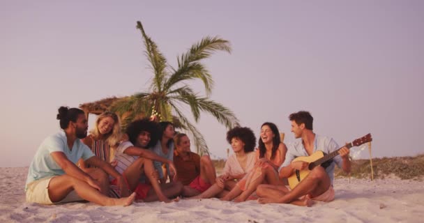 休日の友人の多民族グループのフロントビュー日没で熱帯のビーチで一緒に時間を楽しんで ギターと歌を演奏し スローモーションで — ストック動画