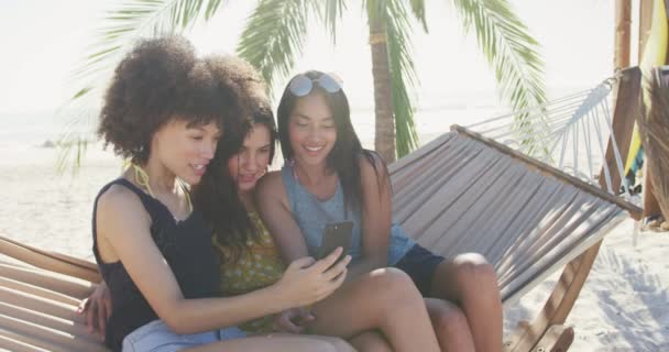 熱帯のビーチで太陽の下で時間を楽しむ友人の多民族グループのフロントビュー ハンモックに座って 笑顔と電話で何かを見て スローモーションで — ストック動画