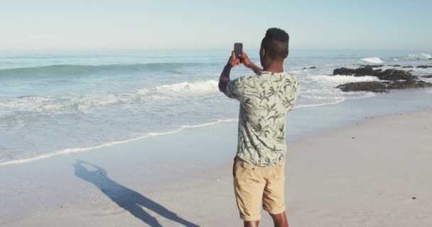 熱帯のビーチで太陽の下で時間を楽しんでいるアフリカ系アメリカ人の男性の後部ビュー 海のパノラマ写真を撮る スローモーションで — ストック動画