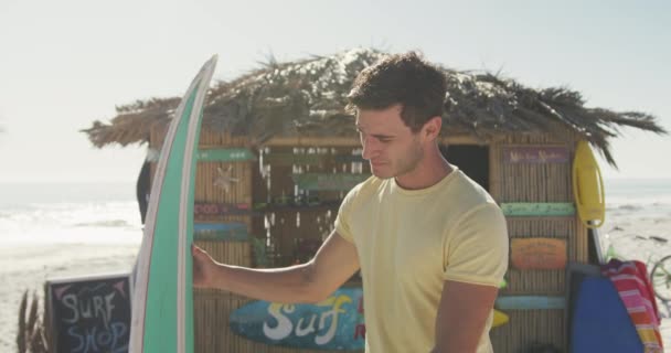 熱帯のビーチで太陽の下で時間を楽しんでいる白人男性のフロントビュー 笑顔と彼の緑のサーフボードを保持 スローモーションで — ストック動画