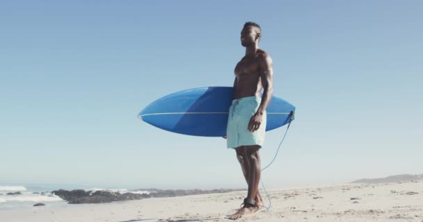 青いサーフボードを持ってゆっくりと動き回るアフリカ系アメリカ人の男が熱帯のビーチで太陽の下で時間を楽しんでいる様子 — ストック動画