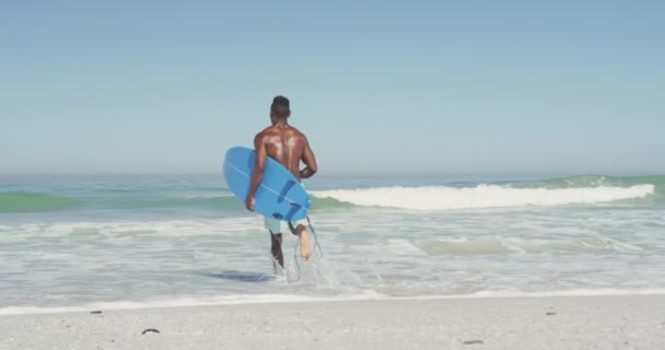 熱帯のビーチで太陽の下で時間を楽しんでいるアフリカ系アメリカ人の男の後ろのビューは 実行し ゆっくりと動きながら 彼の青いサーフボードを運ぶ — ストック動画