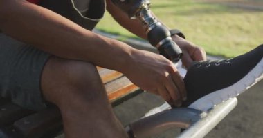 Engelli, protez bacaklı, parkta çalışan, bankta oturan, ayakkabısını bağlayan, kulaklıkla müzik dinleyen bir adamın yan görüntüsü.