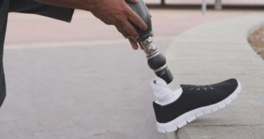 Engelli, protez bacaklı bir ırkın yan görüntüsü. Şehir merkezinde çalışıyor, protez bacağını ayarlıyor, kulaklıkla, ağır çekimde müzik dinliyor.