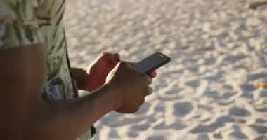Afrika kökenli Amerikalı bir adamın, tropik bir plajda güneşin tadını çıkarırken cep telefonunu kullanıp, yavaş çekimde daktilo kullanmasının yan görüntüsü.