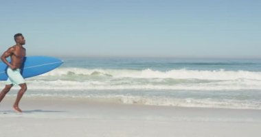 Afrika kökenli Amerikalı bir adamın tropik bir plajda güneşin tadını çıkarırken mavi sörf tahtasını ağır çekimde taşırken ve koşarken yan görüntüsü.