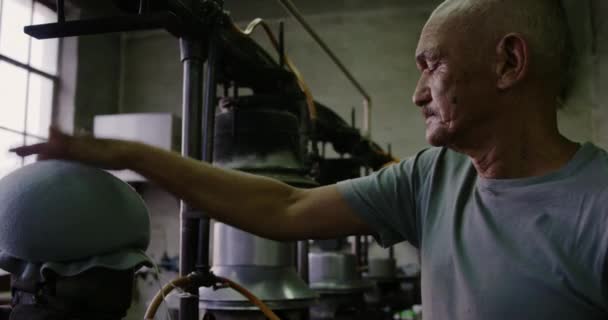 Vue latérale rapprochée d'un homme de race mixte et d'un homme de race mixte senior formant un chapeau dans l'atelier d'une usine de chapeaux, avec des équipements en arrière-plan, au ralenti 