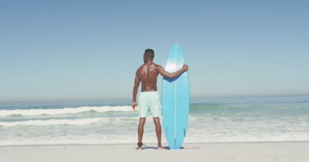 熱帯のビーチで太陽の下で時間を楽しんでいるアフリカ系アメリカ人の男性の後ろのビューは 離れて見て スローモーションで彼の青いサーフボードを保持 — ストック動画