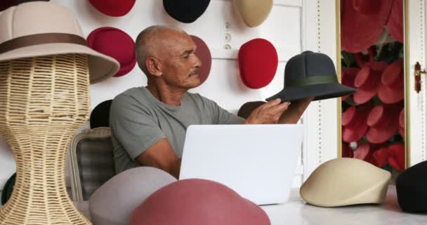 ラップトップコンピュータを使って机の上に座って帽子工場のショールームで完成した帽子を検査して背景に様々な帽子が表示されているスローモーションで — ストック動画