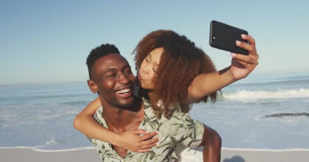 熱帯のビーチで太陽の下で時間を楽しんでいるアフリカ系アメリカ人のカップルのフロントビュー ピギーバック キスをし 自撮り スローモーションで — ストック動画