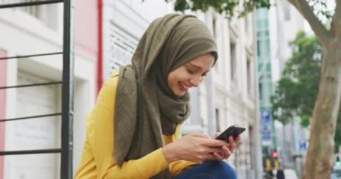 Güneşli bir günde şehirde tesettürlü oturan, akıllı telefon kullanan ve gülümseyen melez bir kadının yan görüntüsü.