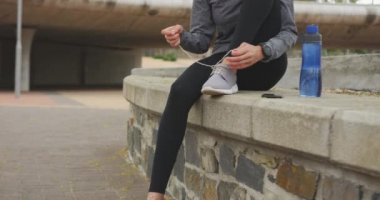 Spor kıyafetleri ve başörtüsü giyen, güneşli bir günde açık havada egzersiz yapan, kulaklık takan, duvarda oturan ve ayakkabılarını yavaş çekimde yanındaki su şişesiyle bağlayan melez bir kadının ön görüntüsü.