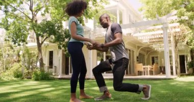 Bahçede eğlenen melez bir çiftin yan görüntüsü. Kadının önünde diz çöküp cebinden yüzüğü çıkaran bir adam, güneşli bir günde, ağır çekimde ona evlenme teklif ediyor.