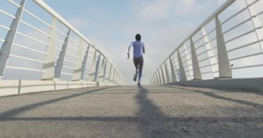 Spor kıyafetleri ve tesettür giyen, güneşli bir günde açık havada mavi gökyüzü ile egzersiz yapan, yavaş çekimde koşan karışık ırktan bir kadının düşük açılı arka görüntüsü.