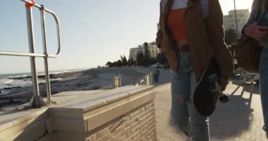 Kafkasyalı ve karışık ırklı bir kızın güneşli bir günde birlikte vakit geçirmesini deniz kenarında bir gezinti alanında ağır çekimde kaykay tutan bir kızın yan görüntüsü..