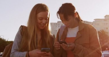 Kafkasyalı ve melez bir kızın güneşli bir günde birlikte vakit geçirmesinden keyif alan, ayakta duran, akıllı telefonlarına bakan, ağır çekimde.