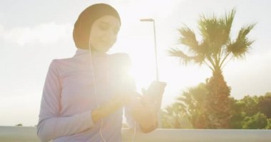 Spor kıyafetleri ve başörtüsü giyen, güneşli bir günde açık havada mavi gökyüzü ile egzersiz yapan, akıllı telefon kullanan ve kulaklık takan, arka planda, yavaş çekimde çalışan melez bir kadının yan görüntüsü.