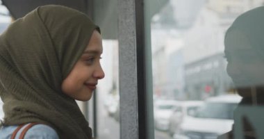 Güneşli bir günde şehirde tesettürlü giyinmiş, yavaş çekimde dükkan camına bakan melez bir kadının yan görüntüsü.