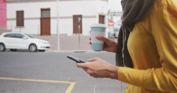 在一个阳光灿烂的城市里 一个身穿头巾的混血儿 拿着外卖咖啡走在大街上 用智能手机慢吞吞地走着 一边看一边看一边看 — 图库视频影像