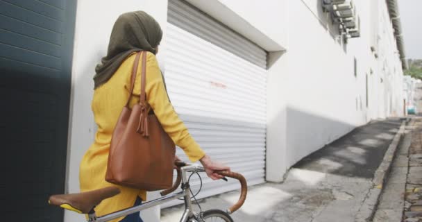 晴れた日に街でヒジャーブを身に着けている混合レースの女性の側面図 通りを歩いて 遅い動きで彼女の自転車をホイール — ストック動画