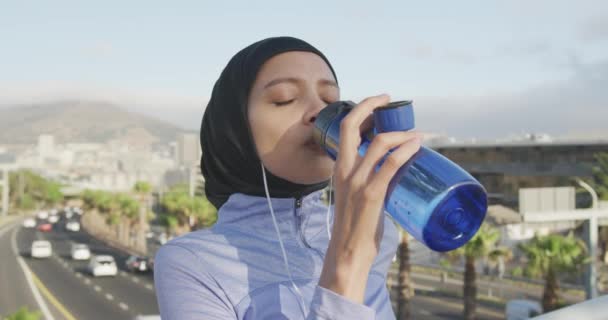스포츠웨어와 히잡을 착용하고 하늘을 배경으로 밖에서 운동을 이어폰을 천천히 마시고 — 비디오