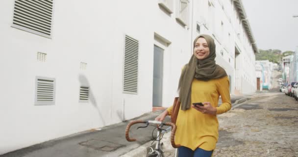 晴れた日に街でヒジャーブを身に着けている混合レースの女性のフロントビュー 通りを歩いて 彼女の自転車をホイール 笑顔とスローモーションでスマートフォンを使用して — ストック動画