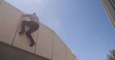 Kafkasyalı bir adam günlük kıyafetler giyiyor güneşli bir günde şehir sokaklarında parkur yapıyor, duvara tırmanıyor ve yavaş çekimde aşağı atlıyor..