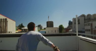 Kafkasyalı bir adam, günlük kıyafetler giyiyor, güneşli bir günde çatıda parkur yapıyor, yavaş çekimde koşuyor ve zıplıyor..