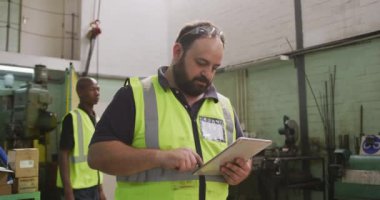 Bir fabrikada çalışan, hidrolik ekipman üreten beyaz bir erkek fabrika işçisi, yüksek bir yelek giyiyor, atölyede duruyor, bilgisayar tabletini kullanıyor, arka planda başka bir işçi geçiyor, yavaş çekte..