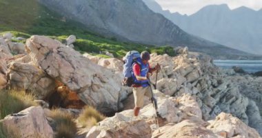 Formda, sakat, protez bacaklı bir ırk adamı, dağlara seyahatinin tadını çıkarıyor, deniz kenarında kayalarda yürüyor, güneşli bir günde, yavaş çekimde, İskandinav bastonları kullanıyor.