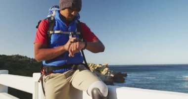 Formda, sakat, protez bacaklı bir ırk adamı, dağlara seyahatinin tadını çıkarıyor, deniz kenarındaki bir bariyerde oturuyor, güneşli bir günde, yavaş çekimde saatini kontrol ediyor.