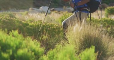 Formda, protez bacaklı, sakat karışık ırk adamı, dağlara seyahatinin tadını çıkarıyor, toprak yolda yürüyor, İskandinav bastonları kullanıyor, güneşli bir günde elinde bir harita tutuyor, yavaş çekimde.