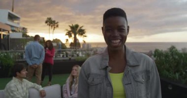 Bulutlu bir akşamda arkadaşlarıyla çatıda takılan ve kameraya ağır çekimde gülümseyen melez bir kadının portresi.