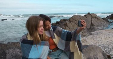 Güneşli bir günde, omuzlarında battaniyeyle sahilde oturmuş, yavaş çekimde selfie çeken, beyaz ırktan bir kızla eğlenen bir kızın yan görüntüsü..