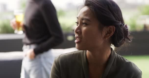 亚洲女人和一个混血男人一起在屋顶的阳台上聊天 他们的朋友喝啤酒 在后台慢吞吞地交谈 — 图库视频影像