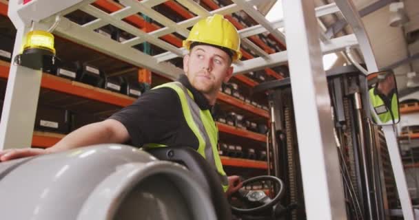 Un ouvrier d'usine de race blanche dans une usine fabriquant des équipements hydrauliques, portant un chapeau dur et une veste haute visibilité, conduisant un chariot élévateur dans une zone de stockage d'entrepôt, au ralenti .