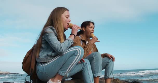 親友同士だ 白人と混じった人種の女の子が晴れた日に一緒に遊びアイスクリームを食べビーチの岩の上に座ってゆっくりとした動きで — ストック動画