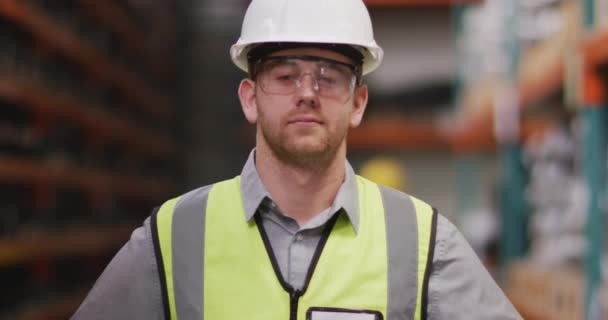 在一家生产液压设备的工厂里 一个白人男性工厂工人头戴硬帽子 戴着安全眼镜 身穿高背心 站在仓库里 慢吞吞地看着镜头 面带微笑 — 图库视频影像